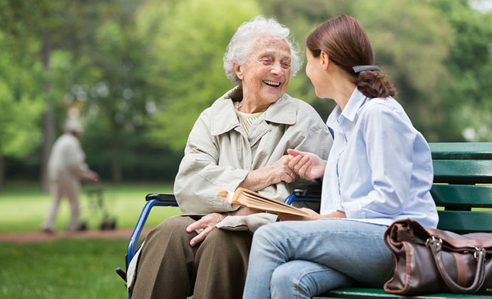 A woman using a wheelchair sits next to and smiles at her daughter, who is seated on a park bench